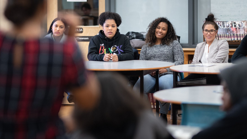 A group of youth sitting a desk listening to a speaker wearing a plaid dress.